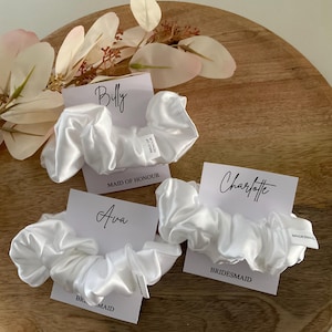 May include: Three white satin scrunchies with personalized cards. The cards have the names "Billy", "Ava", and "Charlotte" and the titles "Maid of Honour" and "Bridesmaid".