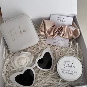 May include: A white gift box containing a selection of items. These include a white jewelry case with "Erika" in gold script, heart-shaped sunglasses, a satin scrunchie, a white rose, and a container with the text "Will you be my bridesmaid?"