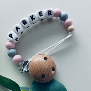 May include: A white silicone baby teether with the name "PARKER" spelled out in black letters. The teether has a wooden clip with three holes and is strung with pink, grey, and beige beads.