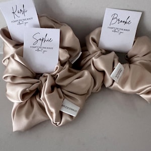 May include: Three beige satin scrunchies with white tags. The tags have the names "Kari", "Sophie", and "Brooke" and the text "I can't tie the knot without you". One scrunchie has a white tag that says "BRIDESMAID".