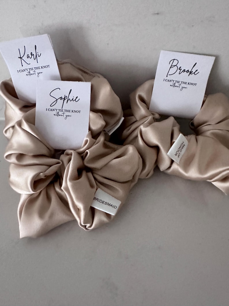 May include: Three beige satin scrunchies with white tags. The tags have the names "Kari", "Sophie", and "Brooke" and the text "I can't tie the knot without you". One scrunchie has a white tag that says "BRIDESMAID".