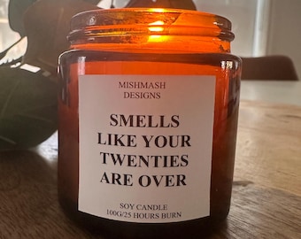 May include: A brown glass jar candle with a white label. The label reads "MISHMASH DESIGNS SMELLS LIKE YOUR TWENTIES ARE OVER SOY CANDLE 100G/25 HOURS BURN". The candle is lit, with a warm orange glow. The candle is on a wooden surface.