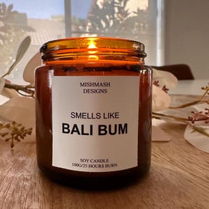 K&ouml;nnte beinhalten: Eine braune Glas-Sojawachskerze mit einem wei&szlig;en Etikett, auf dem "Mishmash Designs", "Smells Like Bali Bum" und "Soy Candle 100g/25 Hours Burn" steht.