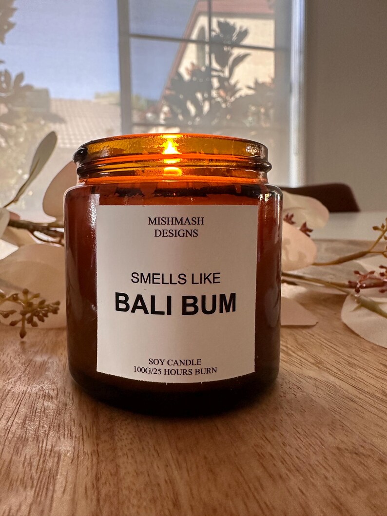 K&ouml;nnte beinhalten: Eine braune Glas-Sojawachskerze mit einem wei&szlig;en Etikett, auf dem "Mishmash Designs", "Smells Like Bali Bum" und "Soy Candle 100g/25 Hours Burn" steht.