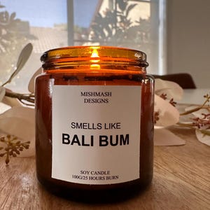 K&ouml;nnte beinhalten: Eine braune Glas-Sojawachskerze mit einem wei&szlig;en Etikett, auf dem "Mishmash Designs", "Smells Like Bali Bum" und "Soy Candle 100g/25 Hours Burn" steht.
