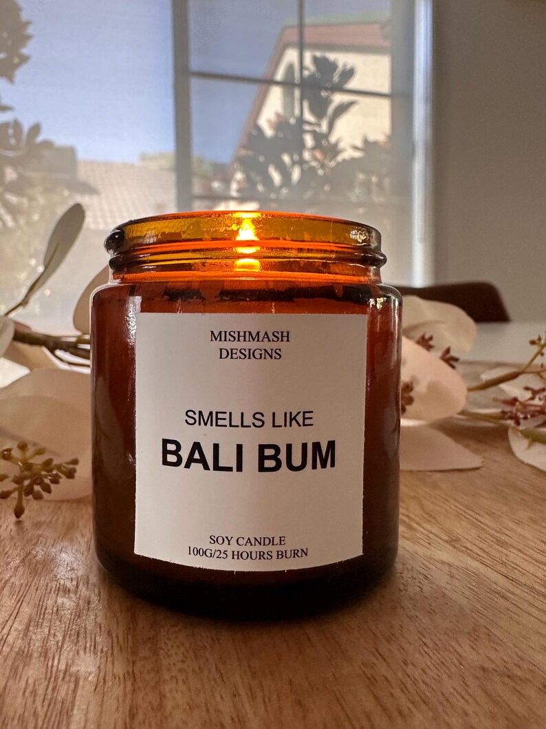 K&ouml;nnte beinhalten: Eine braune Glas-Sojawachskerze mit einem wei&szlig;en Etikett, auf dem "Mishmash Designs", "Smells Like Bali Bum" und "Soy Candle 100g/25 Hours Burn" steht.