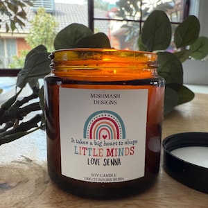 May include: A brown glass jar candle with a black lid. The label features the text "MISHMASH DESIGNS", "It takes a big heart to shape LITTLE MINDS LOVE SIENNA", and "SOY CANDLE 100g/25 HOURS BURN". The candle is a soy candle.