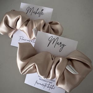 May include: Two beige satin scrunchies with white cards attached. The cards have the text "Michelle" and "Mary" written on them. The card attached to the second scrunchie also has the text "I can't get hitched without my favourite bitch!"