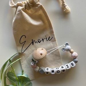 Op de afbeelding: Een gepersonaliseerde baby-bijtring met de naam "Emorie" gespeld in witte kralen met zwarte letters. De bijtring is bevestigd aan een houten clip met een grijze muisbedel en roze en grijze siliconen kralen.