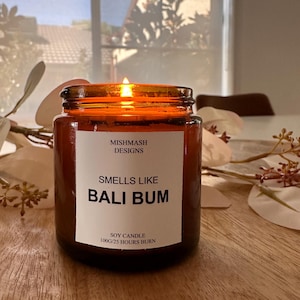 K&ouml;nnte beinhalten: Eine braune Glas-Sojawachskerze mit einem wei&szlig;en Etikett, auf dem "Mishmash Designs" und "Smells Like Bali Bum" steht. Das Etikett tr&auml;gt au&szlig;erdem die Aufschrift "Soy Candle 100g/25 Hours Burn". Die Kerze ist angez&uuml;ndet und die Flamme ist sichtbar.