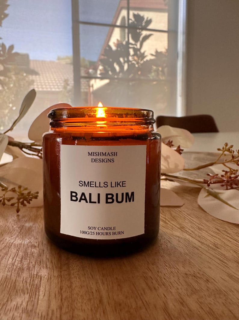 K&ouml;nnte beinhalten: Eine braune Glas-Sojawachskerze mit einem wei&szlig;en Etikett, auf dem "Mishmash Designs" und "Smells Like Bali Bum" steht. Das Etikett tr&auml;gt au&szlig;erdem die Aufschrift "Soy Candle 100g/25 Hours Burn". Die Kerze ist angez&uuml;ndet und die Flamme ist sichtbar.