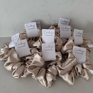 Könnte beinhalten: Neun beigefarbene Satin-Haargummis mit weißen Etiketten, auf denen "Courtney's Hens Weekend" steht.