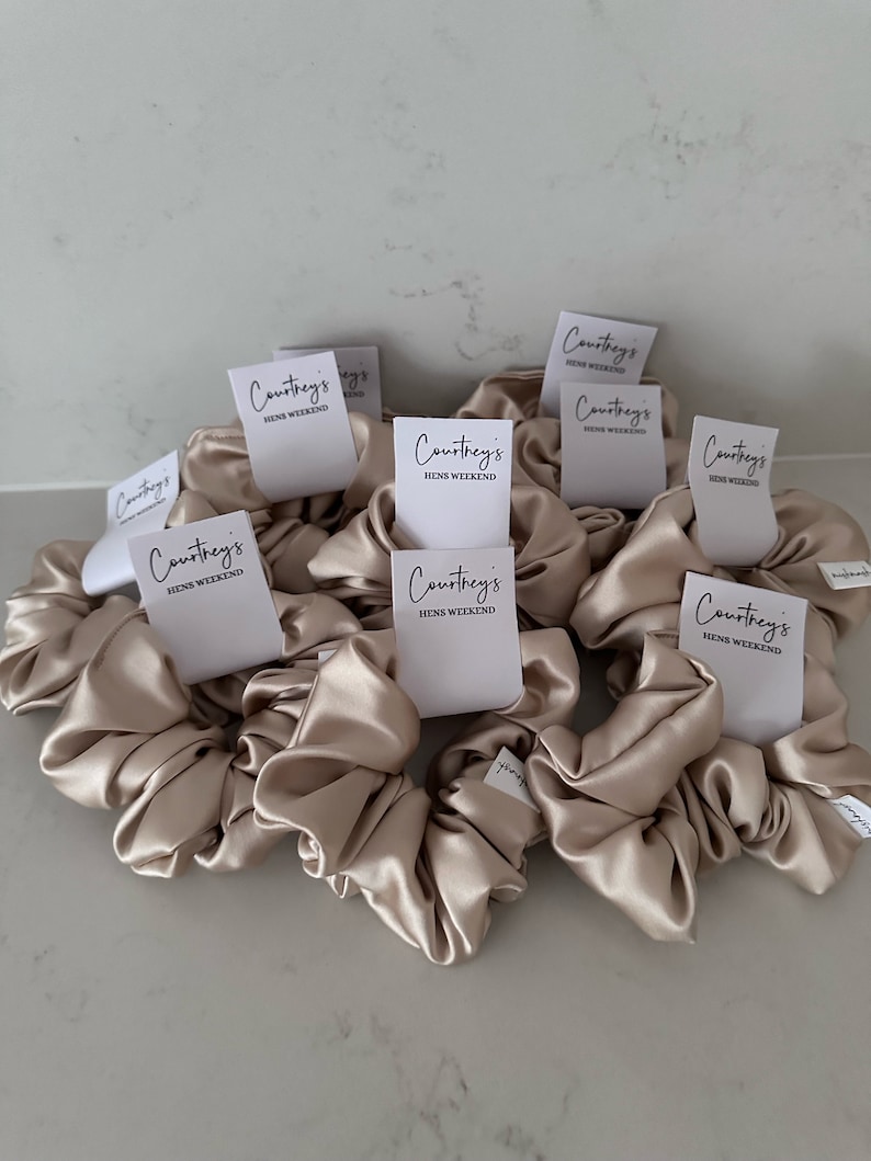 May include: A group of nine beige satin scrunchies with white labels that say "Courtney's Hens Weekend".