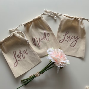 Puede incluir: Tres pequeñas bolsas de tela de color natural con cordón de ajuste, con los nombres "Lara", "Nicole" y "Lucy" escritos en letras doradas rosas. Las bolsas están dispuestas sobre una superficie blanca con un ramo de flores rosas.