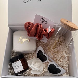 May include: A white gift box with the text "Will You Be Bridesmaid?" contains a scrunchie, a small jewelry case, a candle, heart-shaped sunglasses, a white rose, a glass jar, and a card with the name "Agnes".