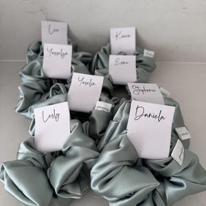 May include: A set of six sage green satin scrunchies with white gift tags attached. The tags have names written on them:  'Liso Yosselin', 'Yoselyn', 'Karina Esme', 'Stephanie', 'Lesly', and 'Daniela'.