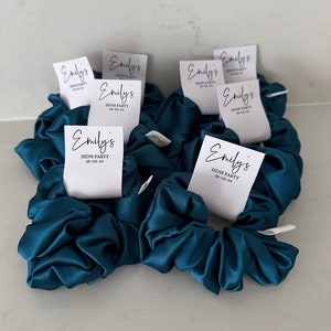 May include: Teal blue satin scrunchies with white labels that say "Emily's Hens Party 25-05-24".