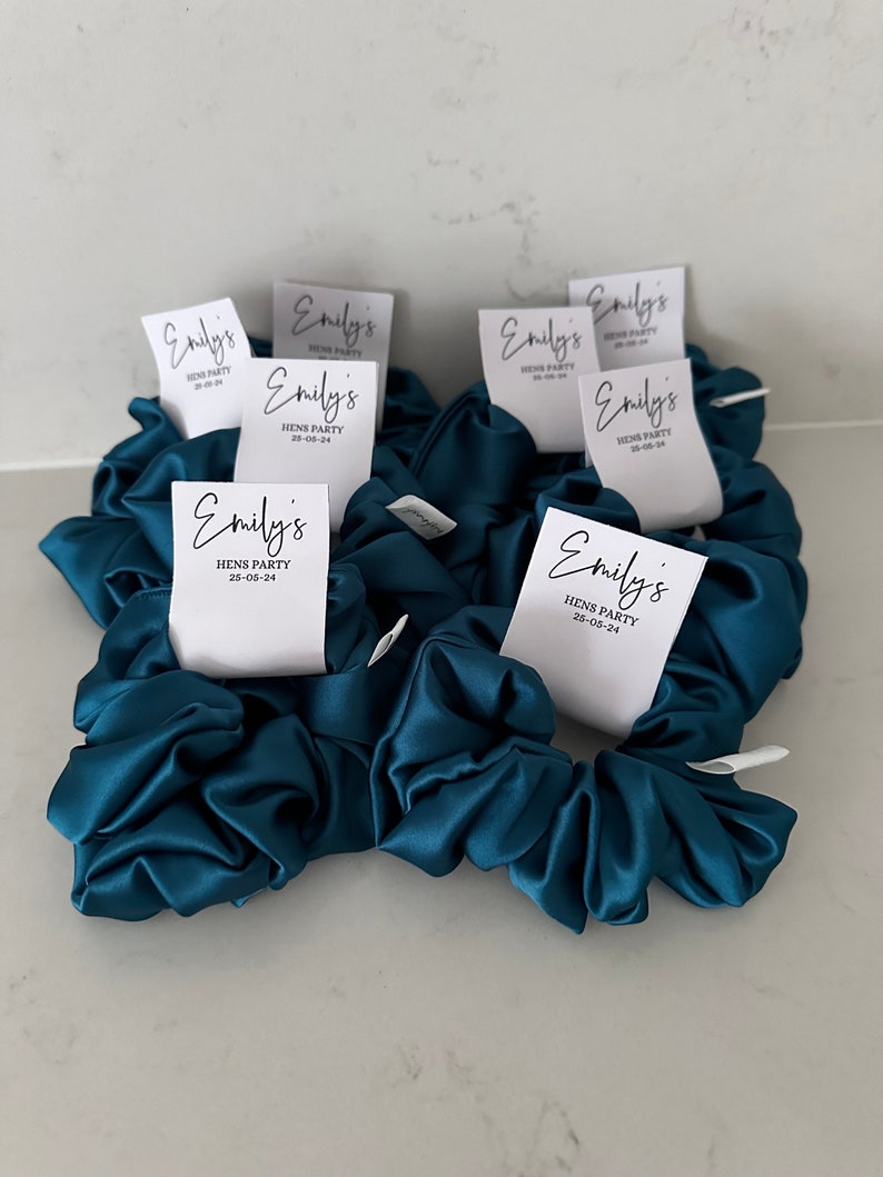 May include: Teal blue satin scrunchies with white labels that say "Emily's Hens Party 25-05-24".