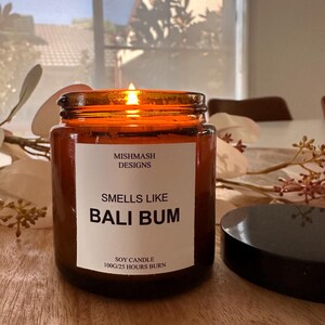 K&ouml;nnte beinhalten: Eine braune Glas-Sojawachskerze mit einem wei&szlig;en Etikett, auf dem "Mishmash Designs, Smells Like Bali Bum, Soy Candle 100g/25 Hours Burn" steht. Die Kerze ist angez&uuml;ndet und die Flamme ist sichtbar.