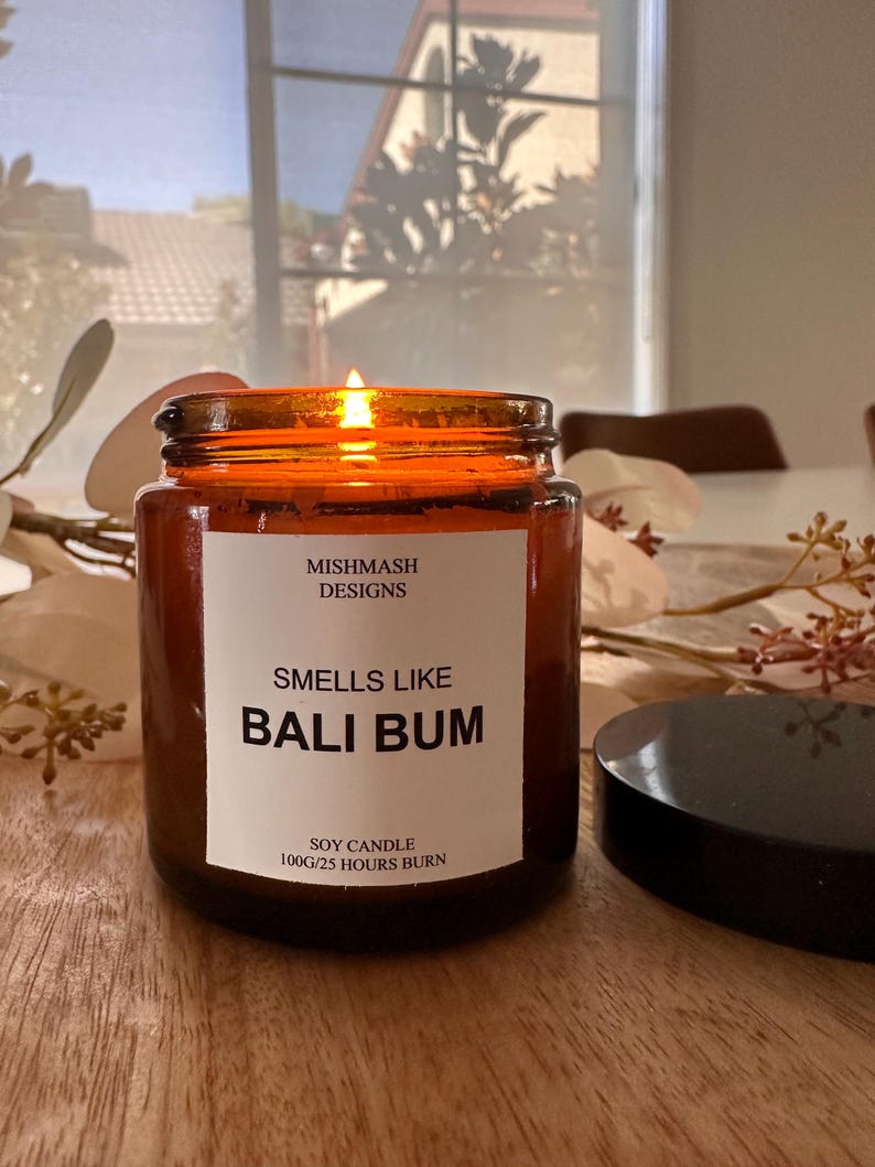 K&ouml;nnte beinhalten: Eine braune Glas-Sojawachskerze mit einem wei&szlig;en Etikett, auf dem "Mishmash Designs, Smells Like Bali Bum, Soy Candle 100g/25 Hours Burn" steht. Die Kerze ist angez&uuml;ndet und die Flamme ist sichtbar.