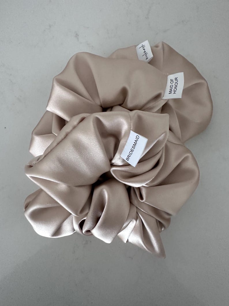 May include: Two satin hair scrunchies in a light beige color. One scrunchie has a tag that says "Bridesmaid" and the other says "Maid of Honor".