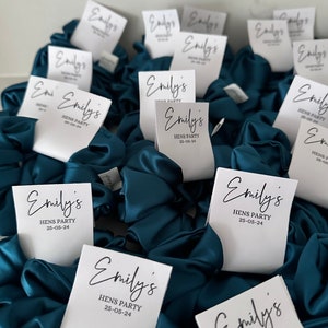 May include: Teal blue satin scrunchies with white labels that say "Emily's Hens Party 25-05-24".