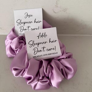 May include: Two purple satin scrunchies with white gift tags. The tags have black text that reads "Josie Sleepover hair Don't care! MILAN" and "Adele Sleepover hair Don't care! MILANA'S 10TH BIRTHDAY".