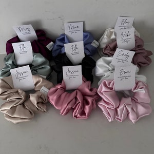 May include: Assortment of satin scrunchies in various colors, each with a personalized card attached. The cards feature different names and messages, such as "Mum", "Ella", "Jessica", and "Erica".