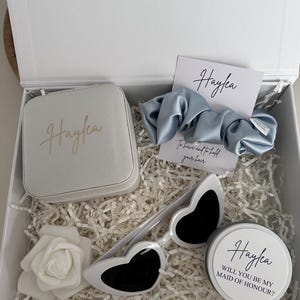 May include: A white gift box filled with items for a bridesmaid. The box contains a white jewelry box with the name "Haylea" on it, a white heart-shaped sunglasses, a white rose, a blue satin scrunchie, and a white round container with the text "Haylea Will you be my Maid of Honour?"