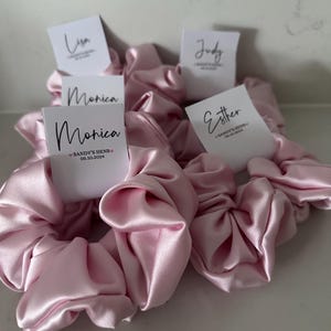 May include: Pink satin scrunchies with personalized name tags. The tags have the names Lisa, Judy, Monica, and Esther. The Monica tag also includes the text "Sandy's Hens 05.10.2024".