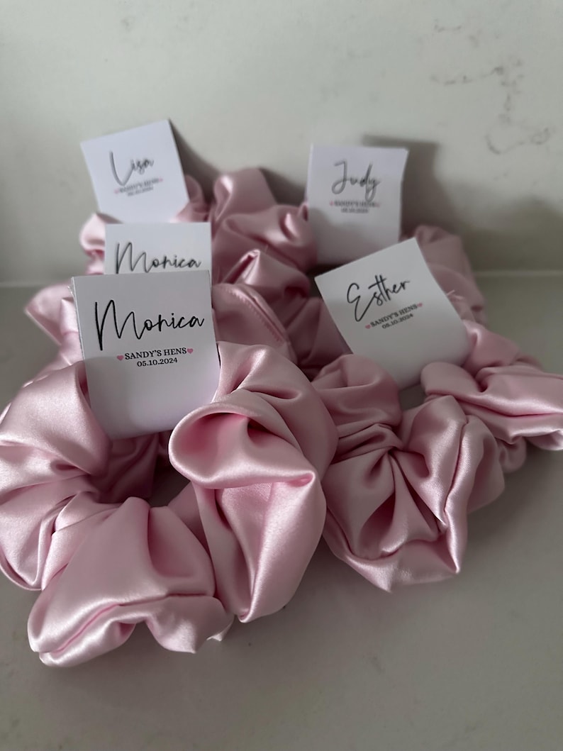 May include: Pink satin scrunchies with personalized name tags. The tags have the names Lisa, Judy, Monica, and Esther. The Monica tag also includes the text "Sandy's Hens 05.10.2024".