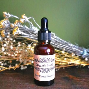 Könnte beinhalten: Eine kleine, bernsteinfarbene Glasflasche mit einem schwarzen Tropfer. Die Flasche hat ein Etikett mit der Aufschrift "Dragon's Blood" und "Holy Roots Botanica".