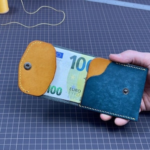 Puede incluir: Cartera de cuero artesanal en color verde azulado y marrón claro, con cierre de botón a presión. Contiene un billete de 100 euros. Se ven herramientas de costura y hilo en el fondo, lo que sugiere un producto hecho a mano.