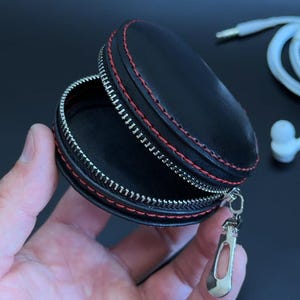 May include: A small, round, black zippered case with red stitching along the edges and a silver metal zipper pull. Beside the case are white wired earbuds with a standard audio jack, designed for storing small electronic accessories.