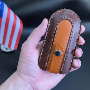 May include: A brown leather key holder with an orange accent and a silver snap closure. The key holder is oval-shaped and has stitched edges. An American flag is in the background.