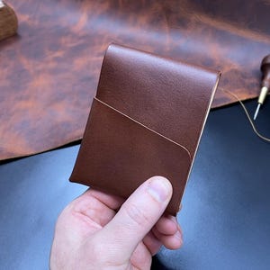 Puede incluir: Cartera de cuero marrón con diseño de bolsillo diagonal. Se ven un cepillo de cuero, monedas y una lata de betún. También hay un carrete de hilo y una herramienta de trabajo del cuero.