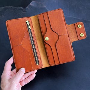May include: Open, brown leather wallet with a zipper pocket and multiple card slots. The wallet is held open, showcasing its interior design and construction. The wallet has a snap closure and is made of high-quality leather.