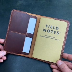 May include: A brown leather notebook cover holding a tan Field Notes memo book and two white cards in internal pockets. The cover features visible stitching along the edges. A separate tan Field Notes memo book and a silver pen are visible nearby.