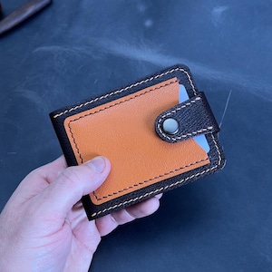 May include: Handmade leather wallet in brown and orange. The wallet has a snap closure and white stitching. It is designed for cards and cash. The background includes leather working tools and a spool of thread. The wallet measures approximately 10 cm x 7 cm.