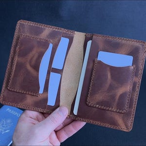 Puede incluir: Portapasaporte de cuero marrón abierto con múltiples ranuras para tarjetas y un compartimento central. El soporte está abierto, revelando inserciones de tarjetas azules claras y una tarjeta blanca. Se ve un pasaporte azul.