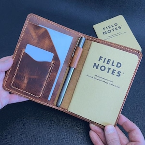 May include: A brown leather notebook cover with a Field Notes memo book and a pen. The cover has card slots and a stitched border. The memo book is tan with the words "FIELD NOTES" printed on the cover. The pen is silver and brown.