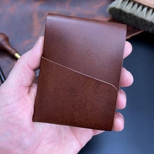 May include: A brown leather cardholder with a front pocket. The rectangular cardholder is likely handmade. The leather has a smooth texture and a rich, warm colour. The background is a blurred leather surface, with a brush visible.