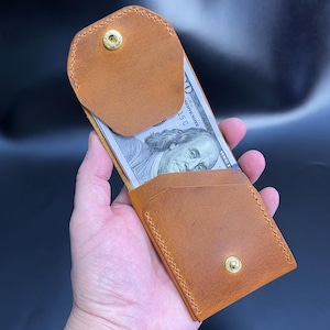 May include: A tan leather wallet with a snap closure, holding a one hundred dollar bill. The wallet is open, revealing the bill. The wallet is held in a hand against a dark background. The wallet is approximately 4 inches long.