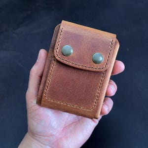 May include: A brown leather wallet with a flap closure secured by two green metal snaps. The wallet is held in a hand, showcasing the textured surface and stitched edges. The overall design is compact and functional.