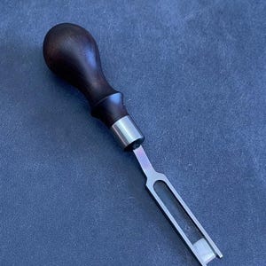 Puede incluir: Una herramienta de carpintería con un mango de madera oscura y una hoja de metal plateado. La hoja tiene un diseño en forma de U con un extremo plano. La herramienta está diseñada para trabajos detallados, probablemente para tallar o dar forma a la madera.