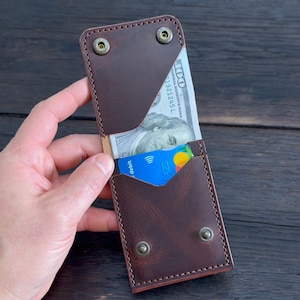 Puede incluir: Una cartera de cuero marrón oscuro con un diseño vertical y costuras visibles en los bordes. La cartera incluye dos botones de presión metálicos en la parte superior y dos en la inferior, conteniendo un billete de cien dólares y una tarjeta.