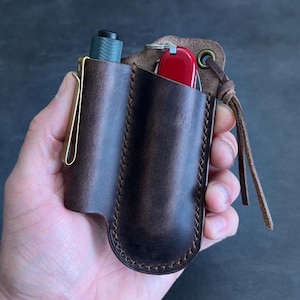 May include: A brown leather multi-tool holder with a brass clip. The holder contains a small flashlight and a red pocket knife. The holder is held in a hand.