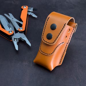 Puede incluir: Una herramienta multiusos con mangos de color naranja brillante y componentes de metal plateado, que incluyen alicates, una hoja y tijeras, junto a una funda de cuero color canela con costuras negras y dos botones a presión negros.
