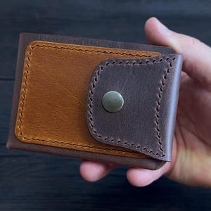 Puede incluir: Una cartera plegable hecha de cuero marrón oscuro y tostado. La cartera presenta un panel rectangular de cuero tostado en la parte frontal, asegurado por una solapa de cuero marrón oscuro con un botón de presión metálico y costuras decorativas.