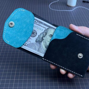Puede incluir: Una cartera de cuero negro hecha a mano con un acento turquesa y costuras blancas. La cartera está abierta, revelando un billete de 100 dólares estadounidenses. La cartera se sostiene en una mano, mostrando su diseño y artesanía.